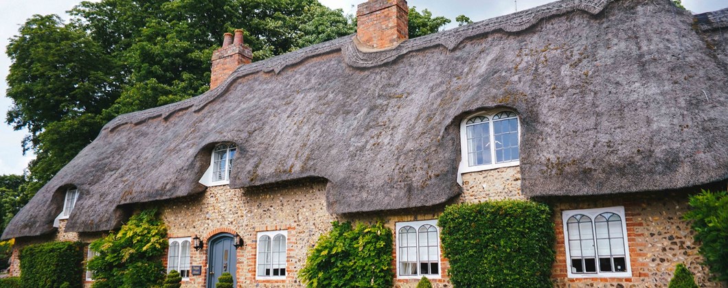 A picturesque cottage with a thatched roof and rustic brickwork, windows and door surrounded by well-tended foliage